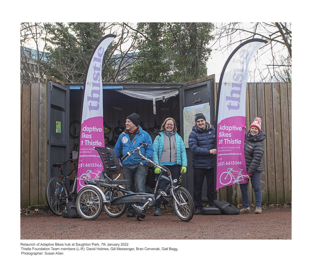 Adaptive bike in front with Thistle charity banners behind and supporters at storage cabin