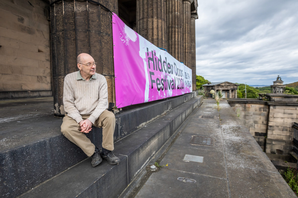 Photo of Derek Grainge a former pupil at the Royal High School sitting near the new-classical columns and beside a Hidden Door banner