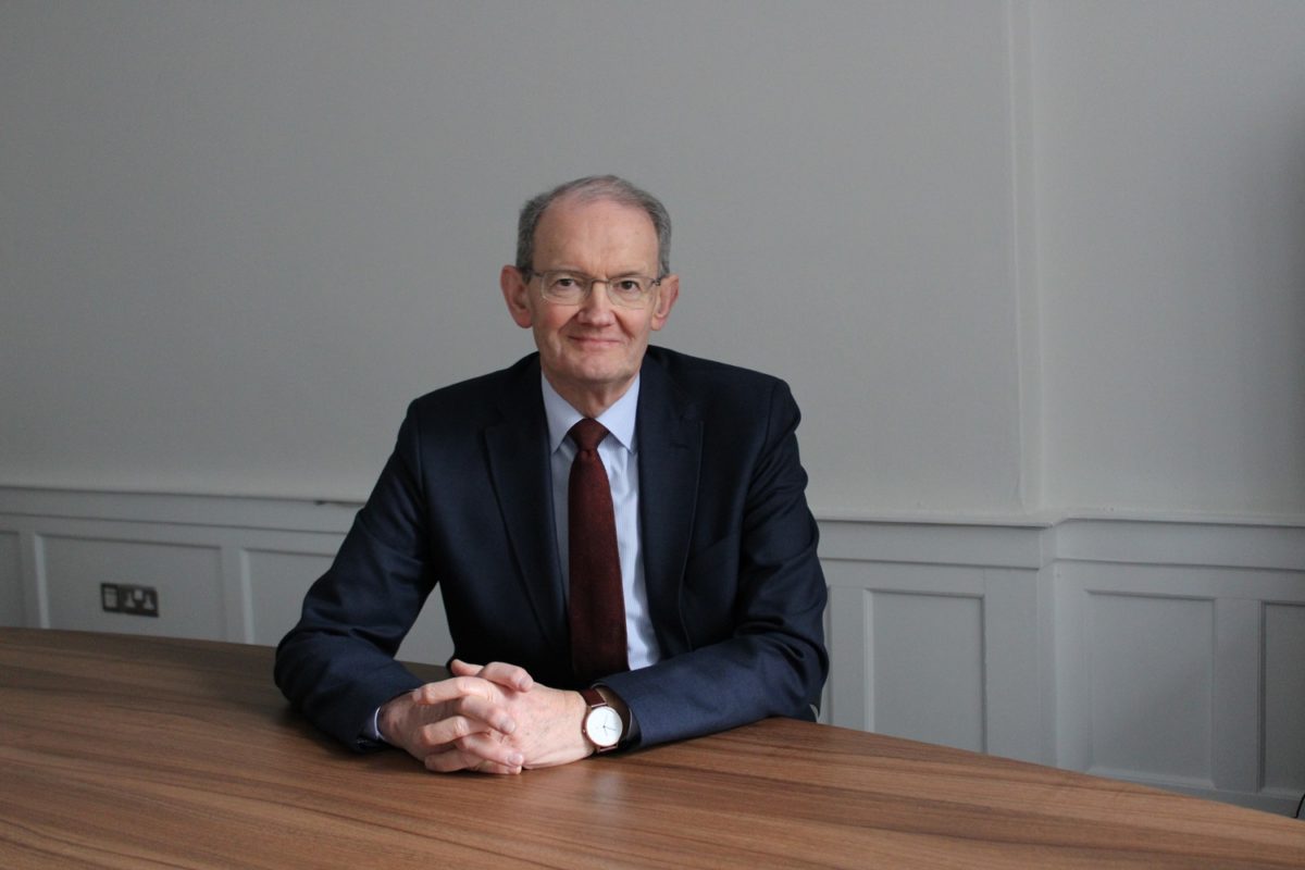 Man in suit sitting at desk with one hand on surface and looking at camera