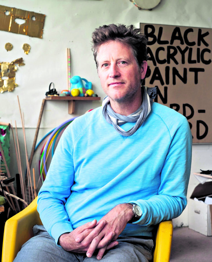 Man in blue sweater sitting on a chair in a workshop with tools on walls looking at camera 