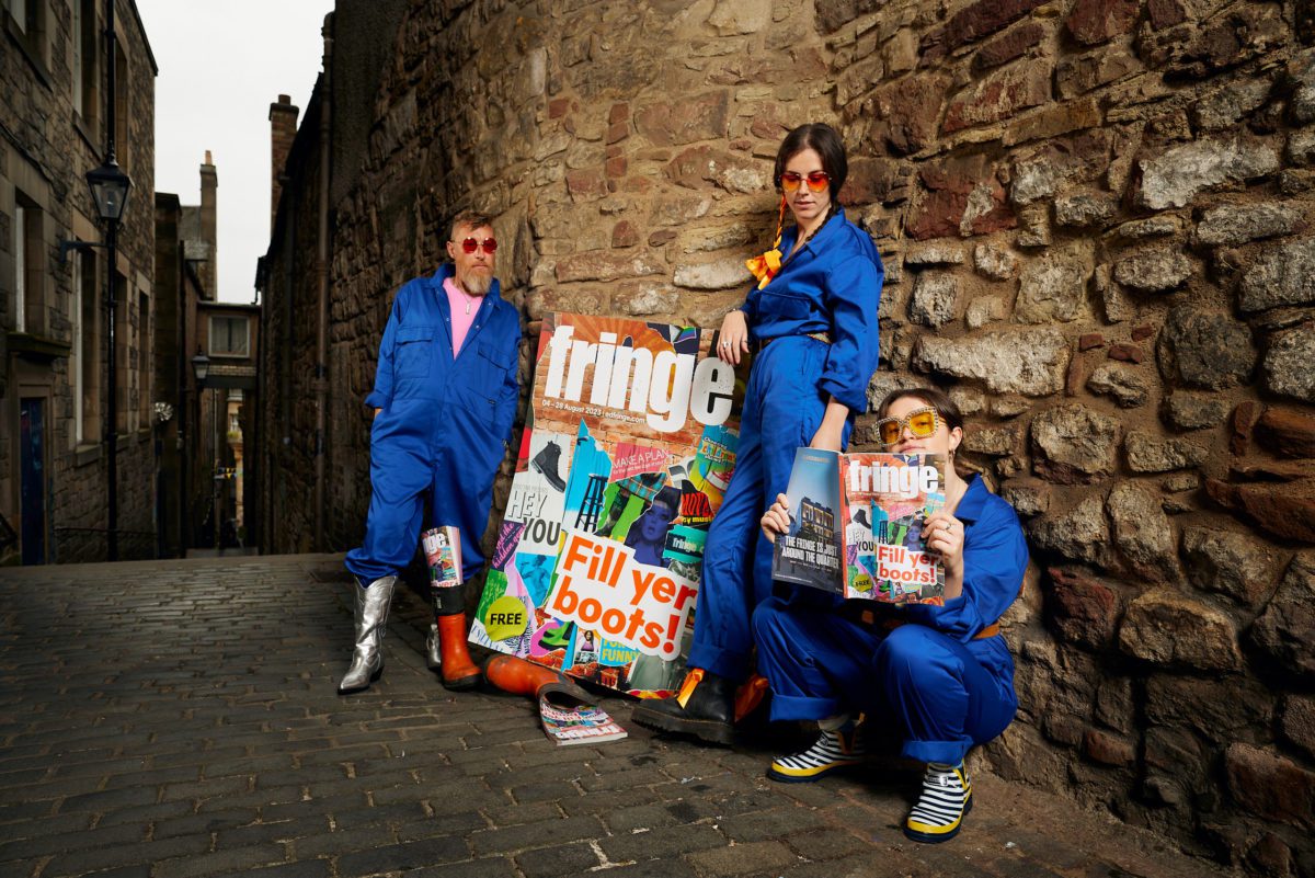 Three people in blue jumpsuits leaning against a brick wall, with a oversized copy of the Fringe 2023 brochure